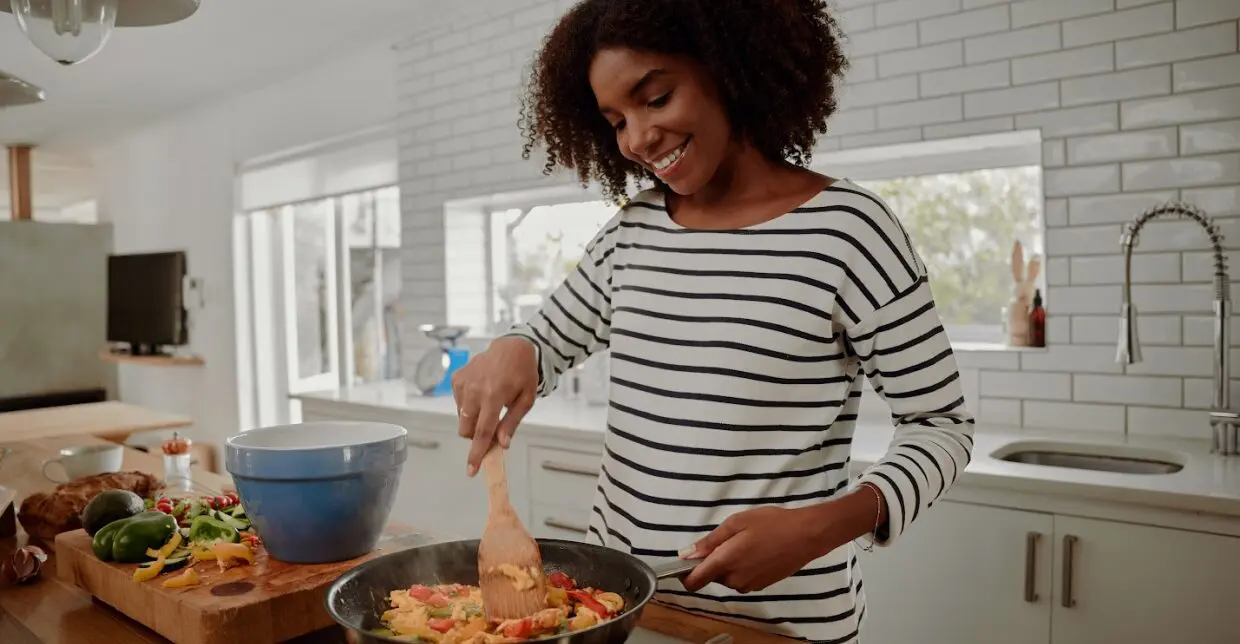 A woman happily cooking in her kitchen, stirring food in a pan.
