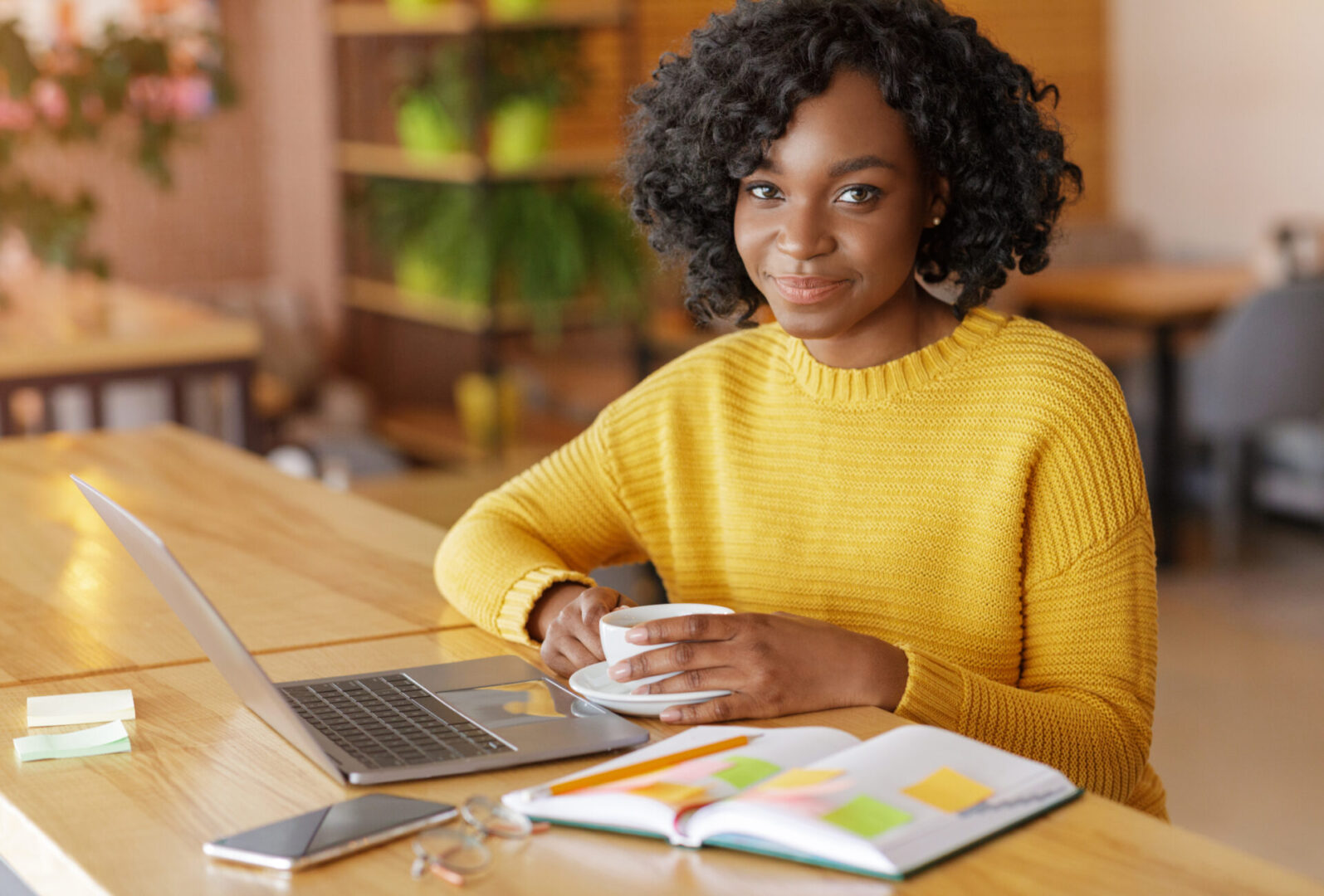 Young woman in a yellow sweater enjoys coffee while working on her laptop.