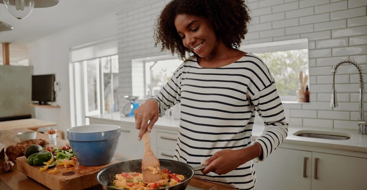 A woman happily cooking in her kitchen, stirring food in a pan.