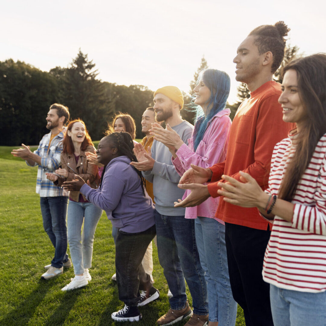 A diverse group of people standing outdoors, clapping and smiling.